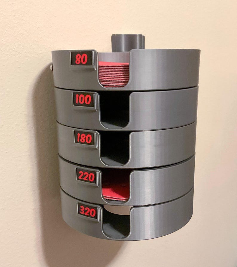 May include: A gray wall-mounted storage organizer for circular sanding discs, featuring five stacked compartments. Each section has a U-shaped cutout and a black label with red text indicating grit levels 80, 100, 180, 220, and 320.