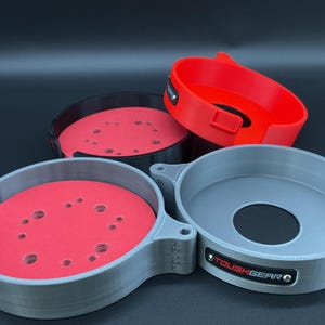 May include: Three circular storage holders for sanding discs, featuring a grey, black, and red color scheme. Each holder has a central opening and a side tab with the text TOUGHGEAR printed on a small black plate attached to the outer rim.