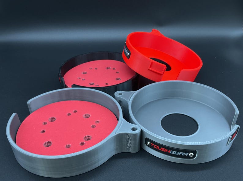 May include: Three circular storage holders for sanding discs, featuring a grey, black, and red color scheme. Each holder has a central opening and a side tab with the text TOUGHGEAR printed on a small black plate attached to the outer rim.