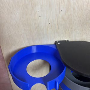 Puede incluir: Un portavasos circular azul con una secci&oacute;n recortada, conectado a una placa de montaje plana negra. Debajo del soporte azul, una estructura cil&iacute;ndrica presenta bandas horizontales alternas en azul y gris con texto visible.