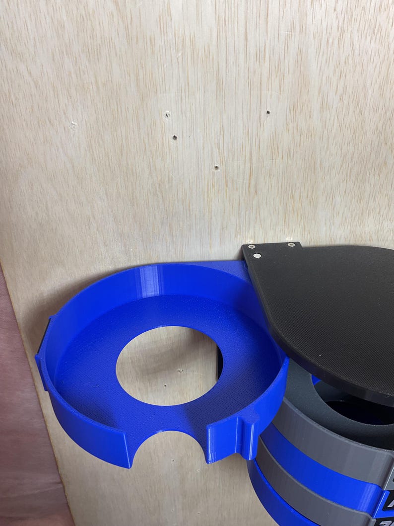 Puede incluir: Un portavasos circular azul con una secci&oacute;n recortada, conectado a una placa de montaje plana negra. Debajo del soporte azul, una estructura cil&iacute;ndrica presenta bandas horizontales alternas en azul y gris con texto visible.