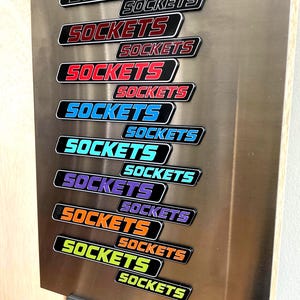 May include: A vertical metal plate featuring multiple rectangular labels, each displaying the word SOCKETS in various colours including white, red, blue, teal, purple, orange, and lime green, arranged in a staggered, overlapping pattern.