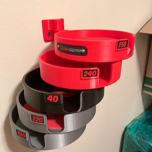 May include: A wall-mounted storage rack featuring five stacked circular holders in red, black, and gray. Each holder is labeled with numerical grit ratings 150, 240, 40, 220, and 320 in red text. The top holder features a black ToughGear logo plate.