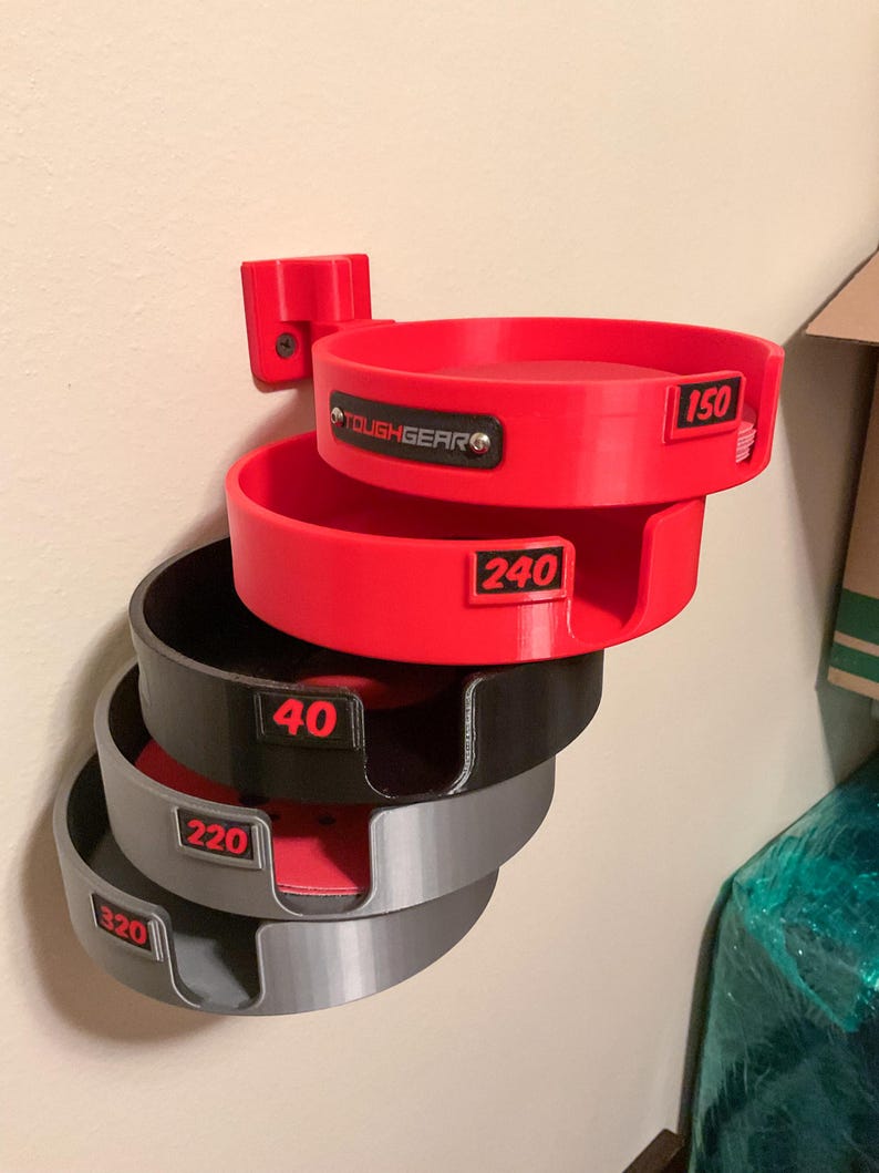 May include: A wall-mounted storage rack featuring five stacked circular holders in red, black, and gray. Each holder is labeled with numerical grit ratings 150, 240, 40, 220, and 320 in red text. The top holder features a black ToughGear logo plate.
