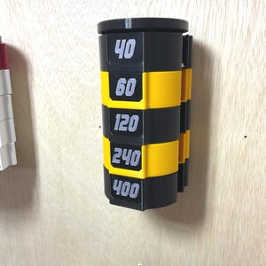 Puede incluir: Un organizador cil&iacute;ndrico montado en la pared con segmentos horizontales que alternan entre negro y amarillo. Las secciones negras tienen los n&uacute;meros blancos 40, 60, 120, 240 y 400, dispuestos verticalmente para indicar el grano de los discos.