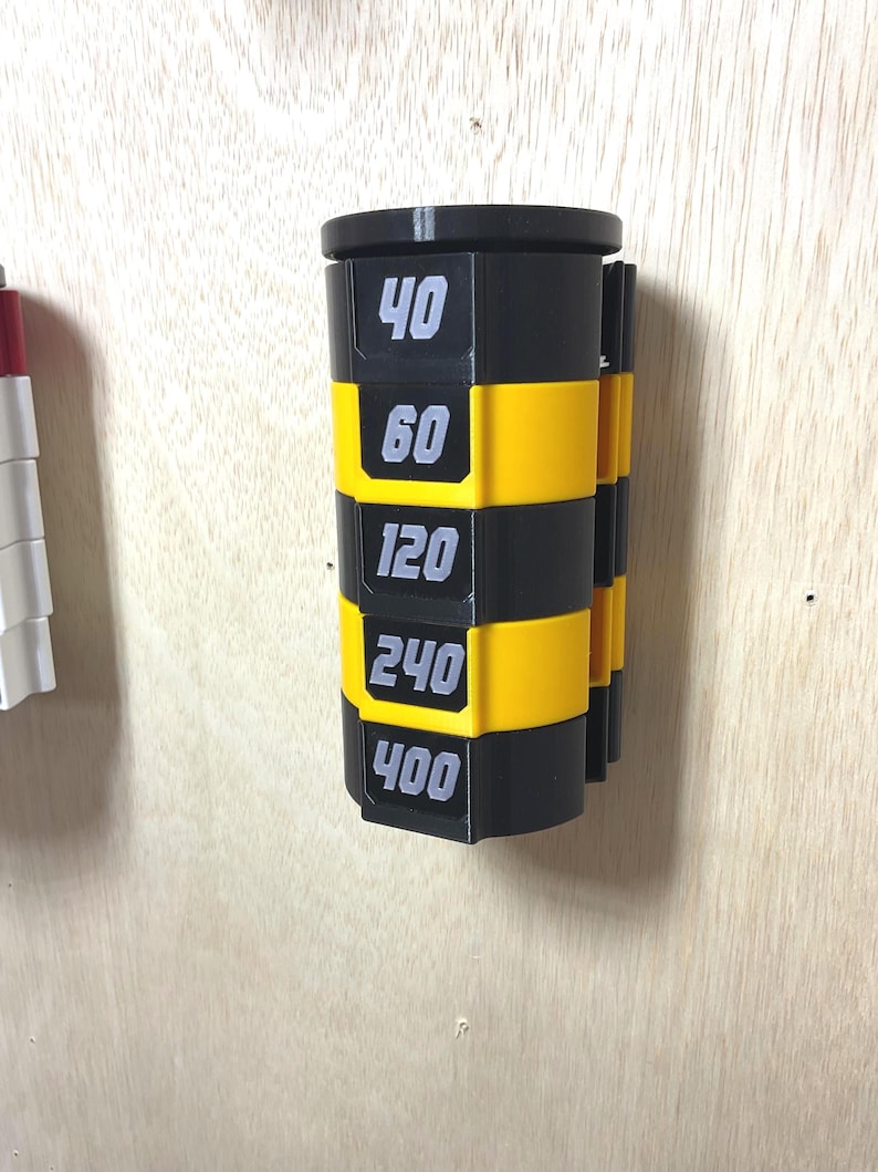 Puede incluir: Un organizador cil&iacute;ndrico montado en la pared con segmentos horizontales que alternan entre negro y amarillo. Las secciones negras tienen los n&uacute;meros blancos 40, 60, 120, 240 y 400, dispuestos verticalmente para indicar el grano de los discos.