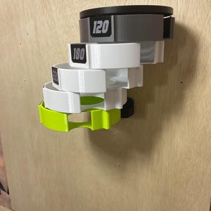 Puede incluir: Un estante de almacenamiento montado en la pared con una pila de cuatro soportes circulares de tama&ntilde;os decrecientes. El soporte superior es negro, seguido de dos soportes blancos y uno verde lima brillante en la parte inferior. Cada soporte muestra una etiqueta num&eacute;rica.