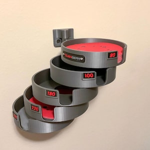 May include: A wall-mounted gray plastic organizer for circular sanding discs, featuring four stacked tiers labeled with red text for grit sizes 80, 100, 180, and 220. The top tier contains a stack of red sanding discs, and the brand name TOUGHGEAR is visible.
