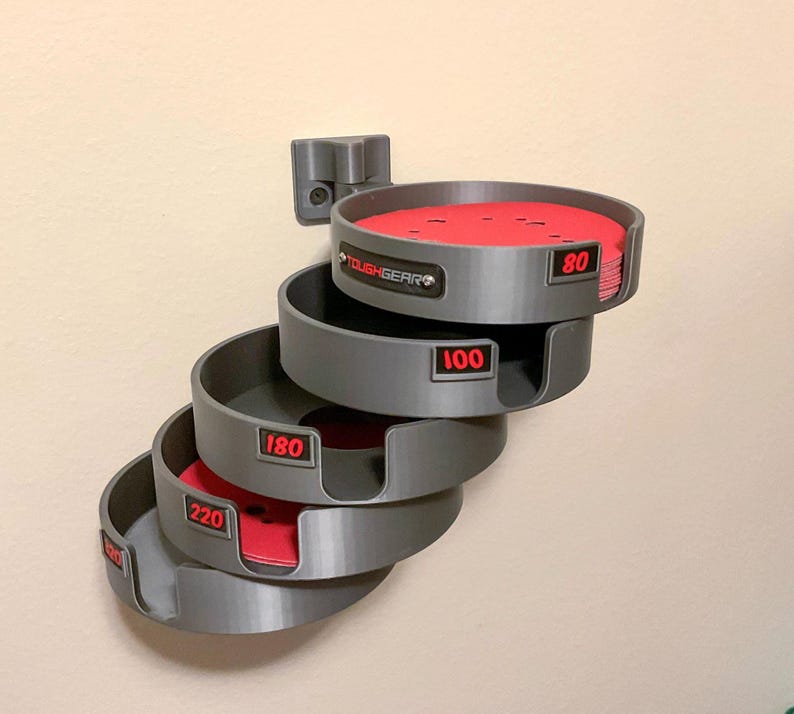 May include: A wall-mounted gray plastic organizer for circular sanding discs, featuring four stacked tiers labeled with red text for grit sizes 80, 100, 180, and 220. The top tier contains a stack of red sanding discs, and the brand name TOUGHGEAR is visible.