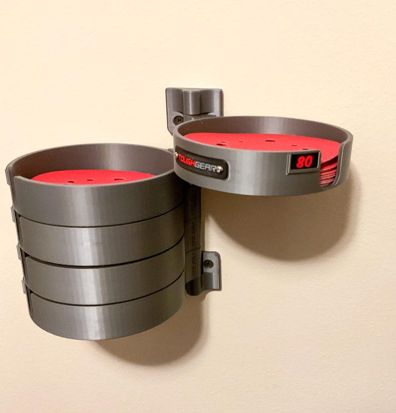 May include: A grey wall-mounted storage organizer with a pivoting arm featuring multiple circular compartments. The unit holds stacks of red circular sanding discs. One compartment displays the text TOUGHGEAR and the number 80 in red.