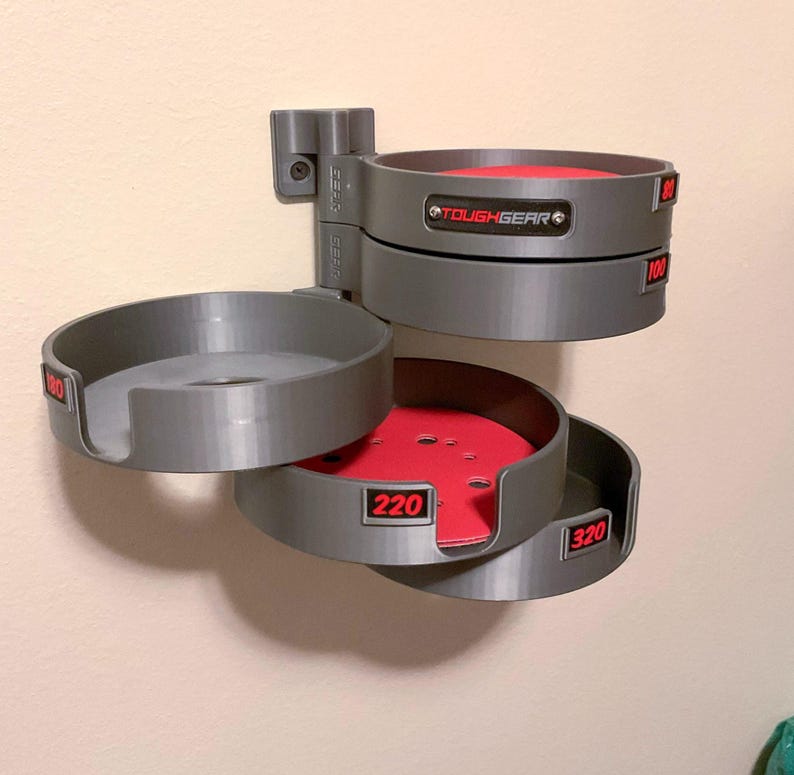May include: A gray wall-mounted organizer with multiple pivoting circular trays for sanding discs. Each tray features a red base and a black label with red text indicating grit sizes 80, 100, 180, 220, and 320. A black plate with the text TOUGHGEAR is attached.