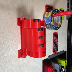 May include: A red wall-mounted organizer featuring five stacked cylindrical holders, each labeled with black numbers 80, 120, 150, 320, and 400. The unit is secured with screws and holds a roll of blue tape on a separate red arm to the right.