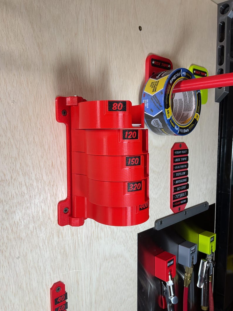 May include: A red wall-mounted organizer featuring five stacked cylindrical holders, each labeled with black numbers 80, 120, 150, 320, and 400. The unit is secured with screws and holds a roll of blue tape on a separate red arm to the right.