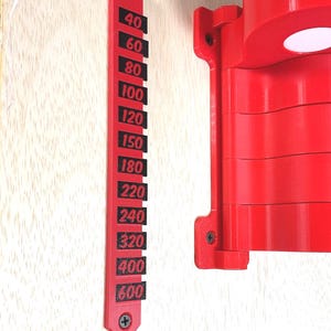 May include: A bright red vertical wall-mounted gauge featuring black rectangular markers with the numbers 40, 60, 80, 100, 120, 150, 180, 220, 240, 320, 400, and 600 printed in red. The gauge is secured with two screws and sits beside a red mechanical component.
