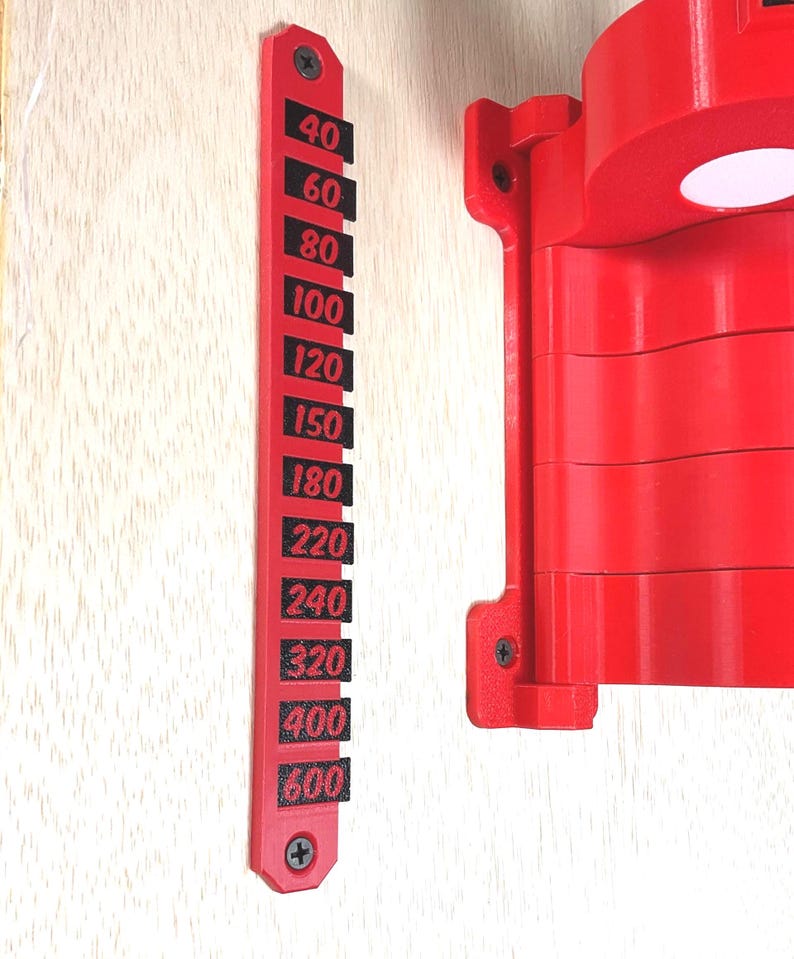 May include: A bright red vertical wall-mounted gauge featuring black rectangular markers with the numbers 40, 60, 80, 100, 120, 150, 180, 220, 240, 320, 400, and 600 printed in red. The gauge is secured with two screws and sits beside a red mechanical component.