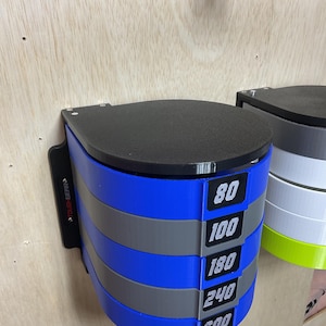 Puede incluir: Un estante de pared para discos de lijado con niveles circulares alternos en azul y gris. Cada nivel tiene una etiqueta negra con texto blanco que muestra los n&uacute;meros 80, 100, 180, 240 y 600. La unidad tiene una tapa negra y el texto TOUGH GEAR impreso en el lateral.