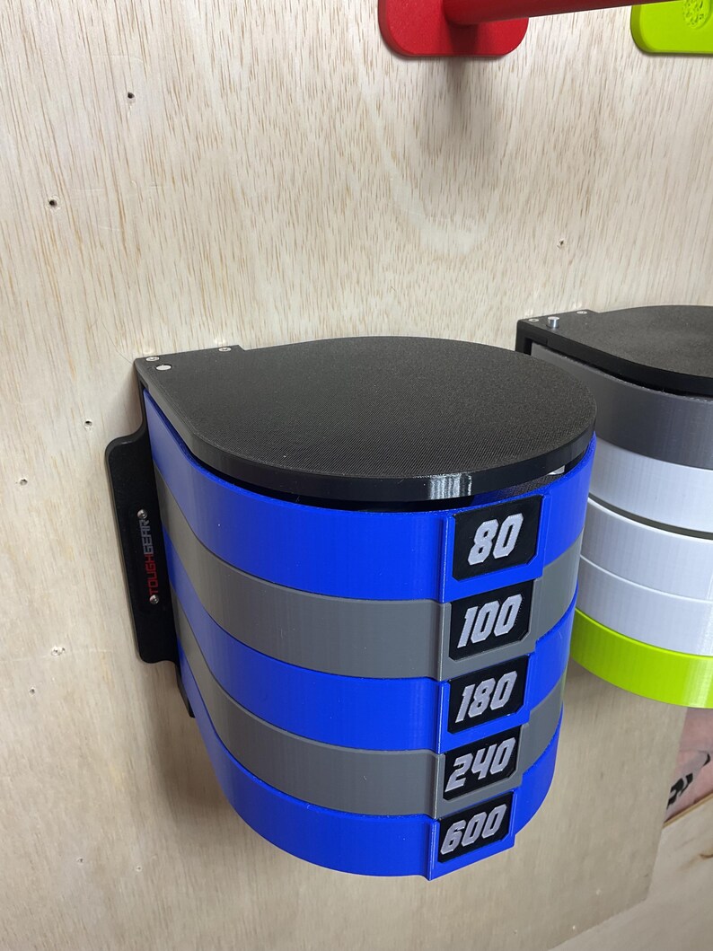 Puede incluir: Un estante de pared para discos de lijado con niveles circulares alternos en azul y gris. Cada nivel tiene una etiqueta negra con texto blanco que muestra los n&uacute;meros 80, 100, 180, 240 y 600. La unidad tiene una tapa negra y el texto TOUGH GEAR impreso en el lateral.