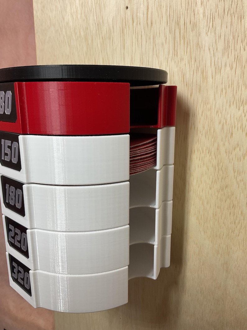 Puede incluir: Un organizador de discos de lija montado en la pared con una parte superior negra y compartimentos de pl&aacute;stico apilados en rojo y blanco. Cada secci&oacute;n est&aacute; etiquetada con los granos 80, 150, 180, 220 y 320 en texto blanco sobre fondo negro.