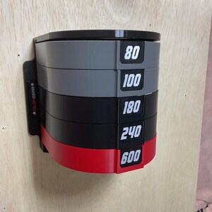 Puede incluir: Un estante de almacenamiento montado en la pared con cinco niveles semicirculares apilados en gris, negro y rojo. Cada nivel muestra una etiqueta num&eacute;rica blanca: 80, 100, 180, 240 y 600. El lateral del soporte negro incluye el texto TOUGHGEAR.