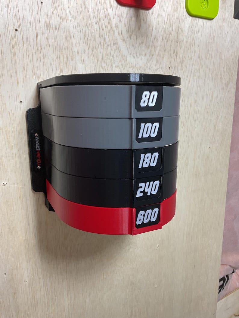 Puede incluir: Un estante de almacenamiento montado en la pared con cinco niveles semicirculares apilados en gris, negro y rojo. Cada nivel muestra una etiqueta num&eacute;rica blanca: 80, 100, 180, 240 y 600. El lateral del soporte negro incluye el texto TOUGHGEAR.