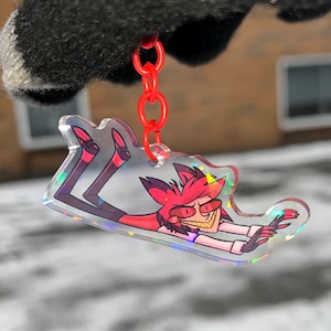 May include: A clear acrylic keychain with a holographic finish featuring a cartoon character with red hair and a pink shirt. The character is lying on their back with their arms outstretched and legs in the air. The keychain is attached to a red chain.