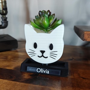 May include: White ceramic cat planter with black eyes and whiskers, holding a small green succulent plant. The planter has a black base with the name "Olivia" written on it.
