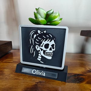 May include: A black and white skull design planter with a faux succulent on top. The planter has a silver frame and sits on a black base with the name "Olivia" on it. The planter is on a wooden surface.