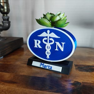 May include: A blue and white Registered Nurse (RN) sign with a Caduceus symbol and the letters "RN". The sign is mounted on a black base with the name "Marta" on it. A green succulent plant sits on top.
