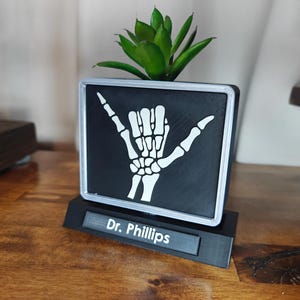 May include: A black and white desktop planter with a skeleton hand design, holding a small green plant. The planter has a silver frame and sits on a black base with the text "Dr. Phillips".