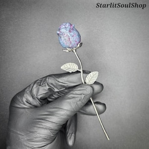 May include: A handcrafted, silver-colored metal rose with a blue and purple stone rosebud. The stem features two detailed leaves. The Etsy shop name "StarlitSoulShop" is in the top right corner.