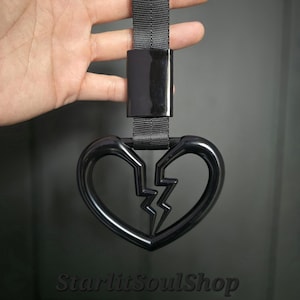 May include: A black heart-shaped accessory with a lightning bolt design, held by a black strap and clasp. The heart is split down the middle. The text "StarlitSoulShop" is at the bottom.