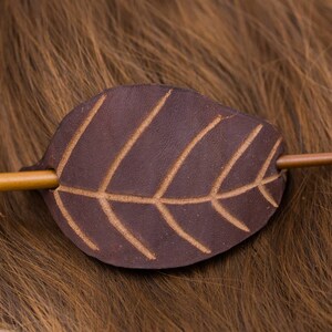 Puede incluir: Una pinza para el pelo de cuero marrón en forma de hoja con un palo de madera adjunto. La pinza está sobre un fondo de pelo marrón.