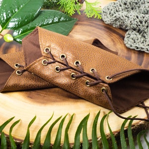 May include: Two brown leather arm warmers with laces and brass eyelets. The arm warmers are on a wooden surface with green leaves.