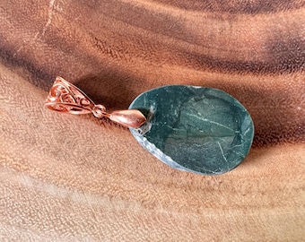 Puede incluir: Un colgante ovalado verde oscuro con una fianza de color cobre. El colgante tiene una superficie lisa y pulida con vetas sutiles.
