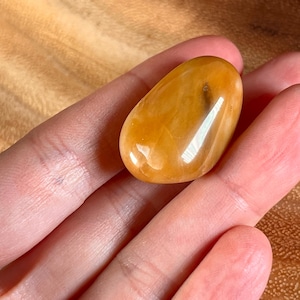 May include: A smooth, polished, orange-colored gemstone with a light brown streak running through it. The gemstone is shaped like a teardrop and is held in a hand.