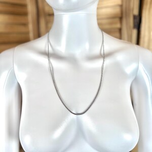 May include: A silver-colored chain necklace displayed on a white mannequin. The chain is a delicate, thin design, and hangs down the mannequin's chest. The background is a light brown wooden surface.