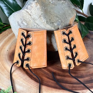 May include: A pair of tan leather wrist cuffs with black laces and brass eyelets. The cuffs are tied together with the laces.