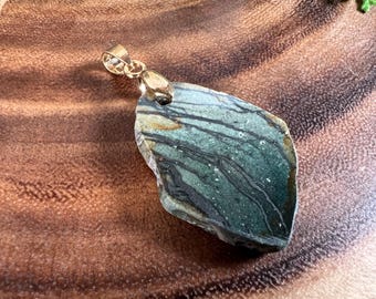 Puede incluir: Un colgante de piedra verde y negra con una anilla de oro. La piedra tiene un patrón único de mármol.
