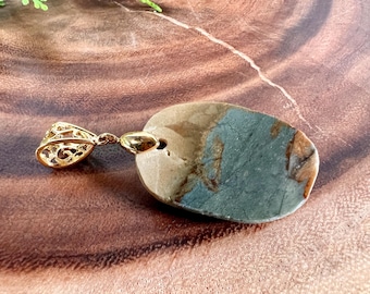 Puede incluir: Un colgante de tono dorado con una piedra ovalada única que presenta una mezcla de colores marrón, verde y gris. La piedra tiene un aspecto natural marmoleado.