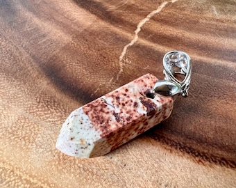 Puede incluir: Un colgante de piedra blanca y marrón con una anilla de plata. La piedra tiene un patrón moteado y tiene forma de rectángulo largo y delgado.