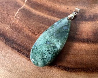 Puede incluir: Un colgante en forma de lágrima hecho de ágata verde musgo con una anilla de plata. La piedra tiene un patrón moteado verde y blanco.