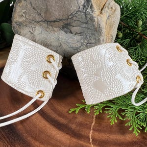 May include: A pair of white lace-up wrist cuffs with gold eyelets. The cuffs have a textured surface and are made of a soft, pliable material.