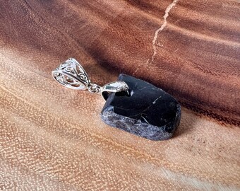 Puede incluir: Un colgante de piedra negra con una argolla de plata. La piedra tiene una forma irregular y una textura rugosa.