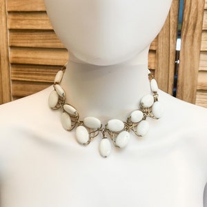 May include: A white oval-shaped beaded necklace with gold wire accents. The necklace is on a white mannequin.