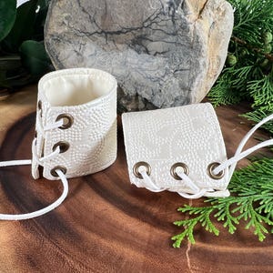 May include: Two white leather wrist cuffs with white laces and brass eyelets. The cuffs are on a wooden surface.