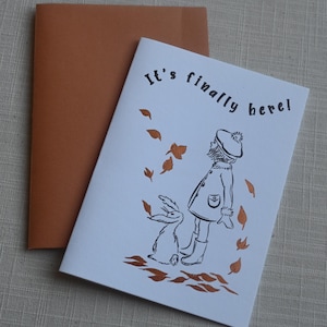 May include: A white greeting card with a brown envelope. The card features a black line drawing of a person in a coat and hat standing with a rabbit, surrounded by copper-coloured falling leaves. The text "It's finally here!" is printed in black.