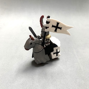 以下が含まれることがあります： 銀色の馬に乗った白黒の騎士のフィギュア。騎士は白いマントを着て剣を持っている。馬は赤いたてがみと尻尾をしている。騎士は黒い十字架のついた白い旗を持っている。
