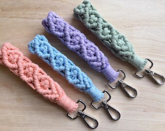 May include: Four colorful macrame keychains with silver key rings. The keychains are made of woven cotton cord in shades of pink, blue, purple, and green.