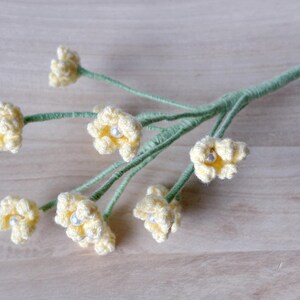 May include: A bunch of crocheted yellow flowers with white pearl centers on green stems. The flowers are arranged in a bouquet and are on a light brown wooden surface.