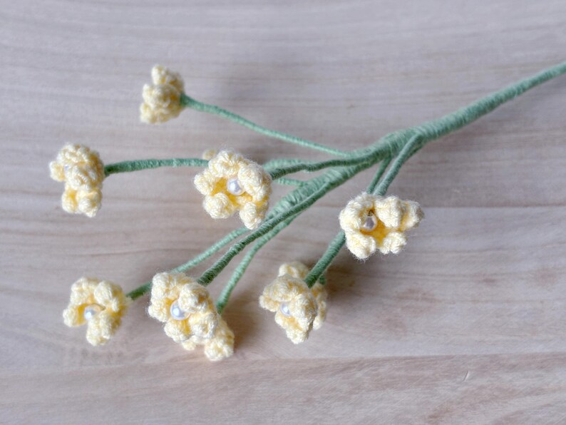 May include: A bunch of crocheted yellow flowers with white pearl centers on green stems. The flowers are arranged in a bouquet and are on a light brown wooden surface.